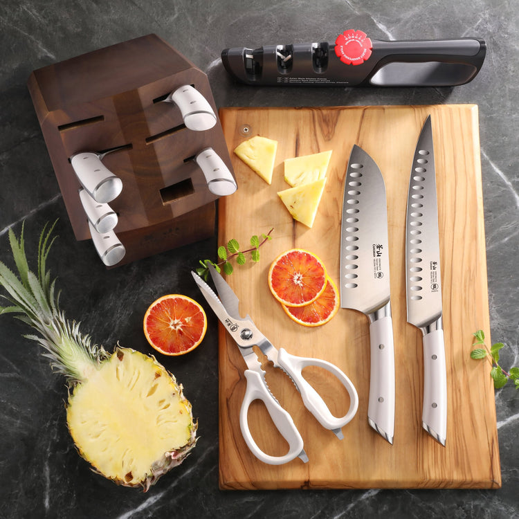 Knife block with six knives removed, two knives, kitchen scissors, knife sharpener, pineapple half, and sliced citrus on wood