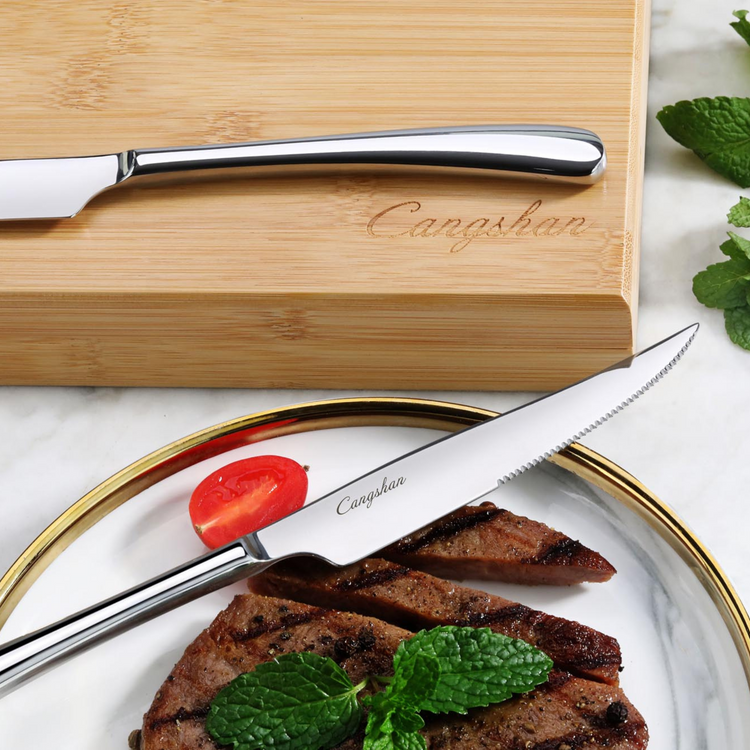 Stainless steel steak knife with serrated edge on a plate with grilled steak, tomato slice, and mint leaves, next to a bamboo
