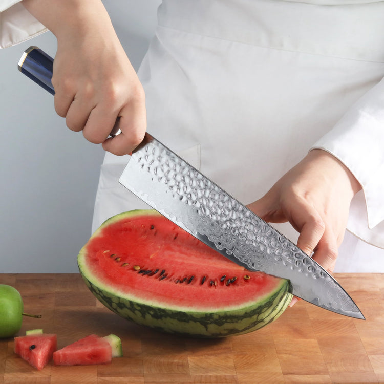 Hand holding a blue-handled chef's knife cutting a watermelon half on a wooden surface.