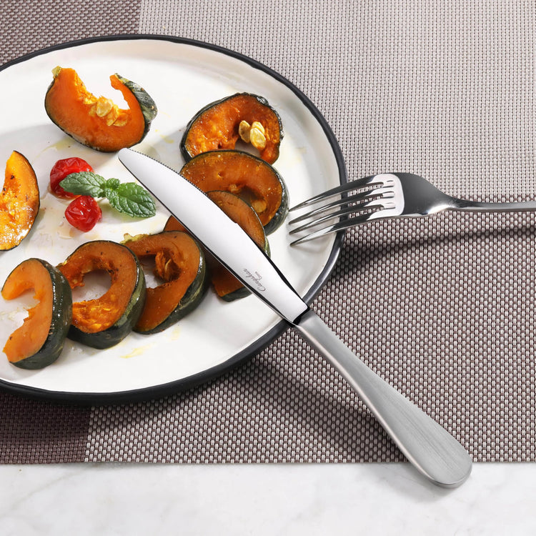 Dinner plate with roasted pumpkin slices, cherry tomatoes, basil, and a stainless steel dinner knife and fork on a placemat.