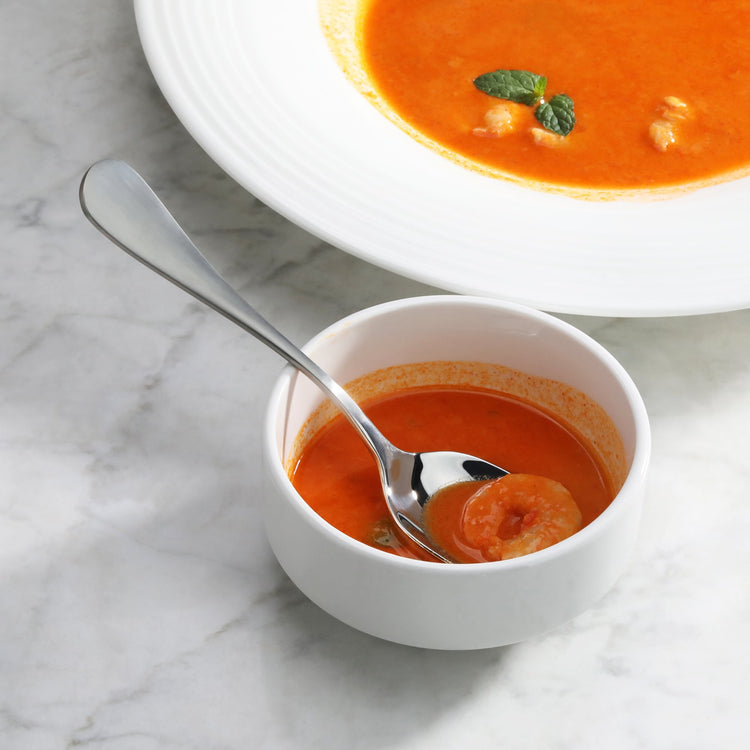 Stainless steel dinner spoon with matte finish handle resting in a white bowl of tomato soup with shrimp.