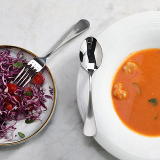 
                    
                      Polished stainless steel dinner spoon with matte finish handle placed on a white plate beside a bowl of orange soup and a sal
                    
                  