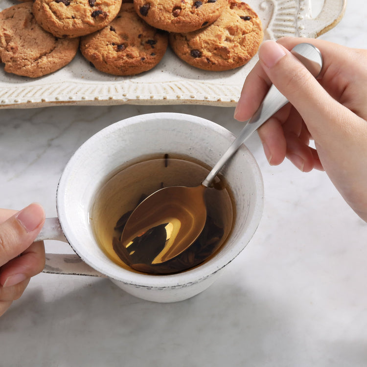 Hand holding a stainless steel teaspoon stirring tea in a white ceramic cup with cookies on a tray nearby.