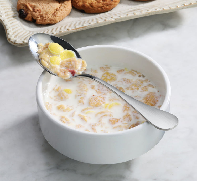 Stainless steel teaspoon with matte finish handles holding cereal and milk above a white bowl of cereal and milk.
