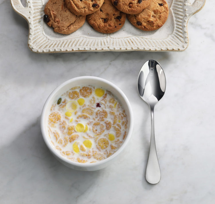 Single stainless steel teaspoon with matte finish handle next to a bowl of cereal and a plate of cookies on a marble surface.