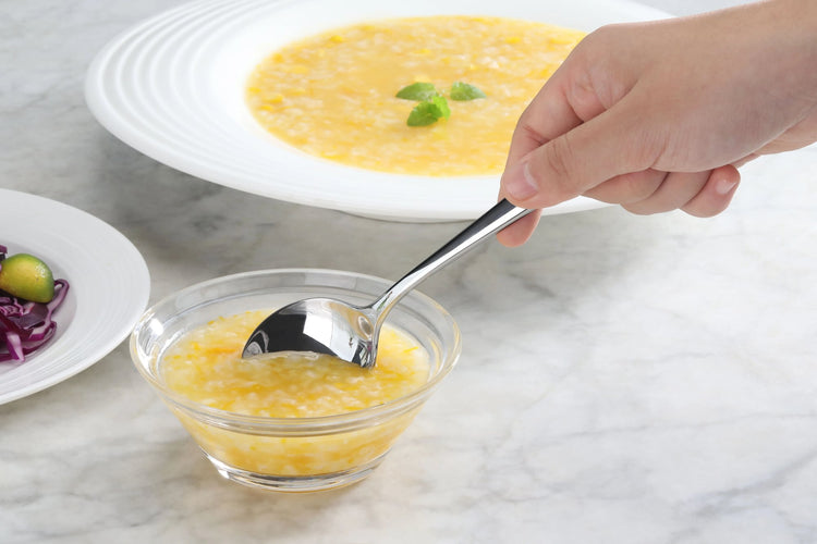 Hand holding a stainless steel spoon scooping yellow soup from a small glass bowl on a marble surface.