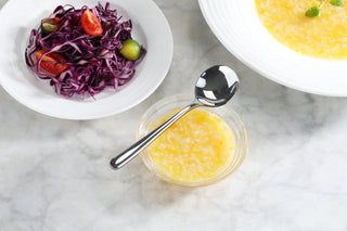 
                    
                      Forged stainless steel bouillon spoon resting on a small bowl of yellow soup, with salad and soup bowls nearby.
                    
                  