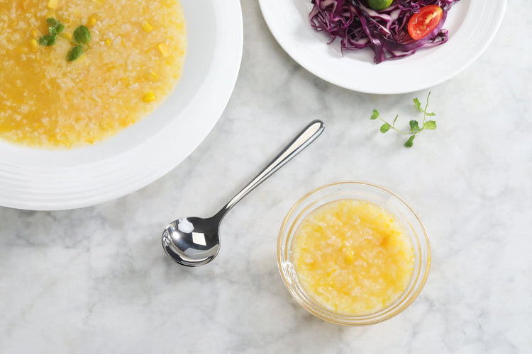 Small stainless steel spoon with a mirror finish placed next to a bowl of yellow soup on a marble surface.