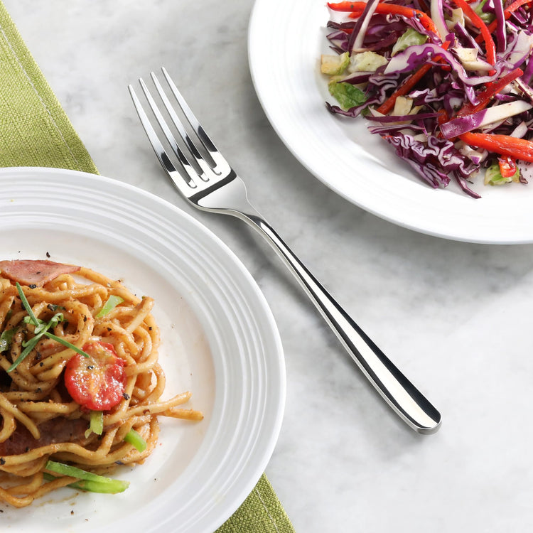 Fork with a mirror finish placed between a plate of noodles and a plate of salad on a marble surface.