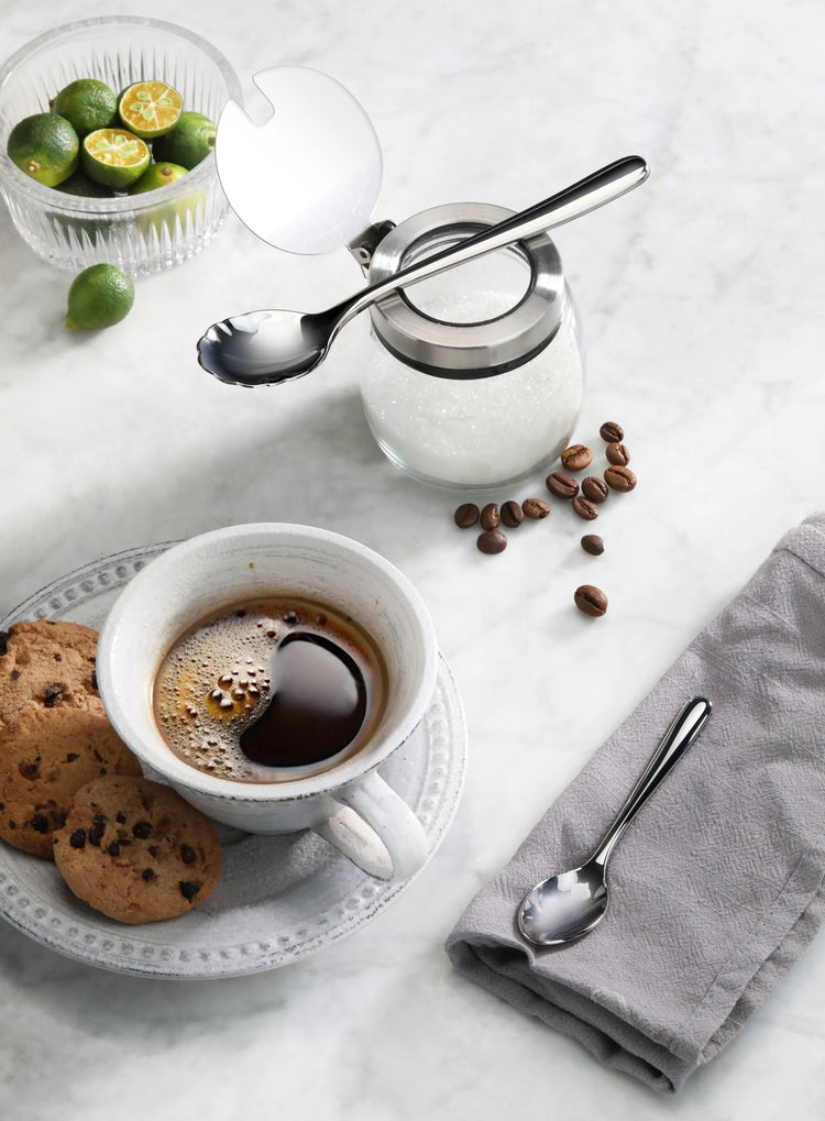 Stainless steel espresso spoon resting on a sugar jar, with coffee cup, cookies, coffee beans, and limes nearby.