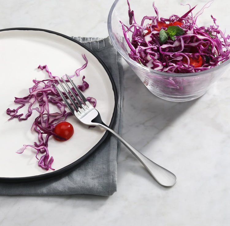 Stainless steel salad fork with matte finish handle on a plate with shredded red cabbage and cherry tomato next to a glass bo