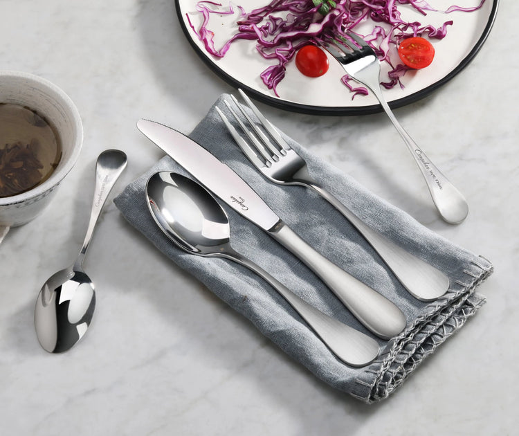 Salad fork, knife, and spoon with matte finish handles on a gray napkin next to a plate with salad and cherry tomatoes.