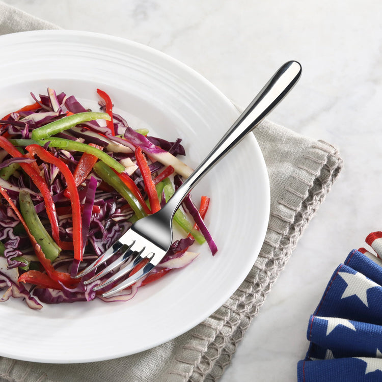 Salad fork with mirror finish resting on a white plate of mixed sliced vegetables on a gray cloth.