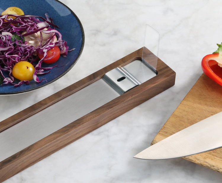 Walnut magnetic knife bar with metal strip on marble countertop next to cutting board, knife, and salad plate.