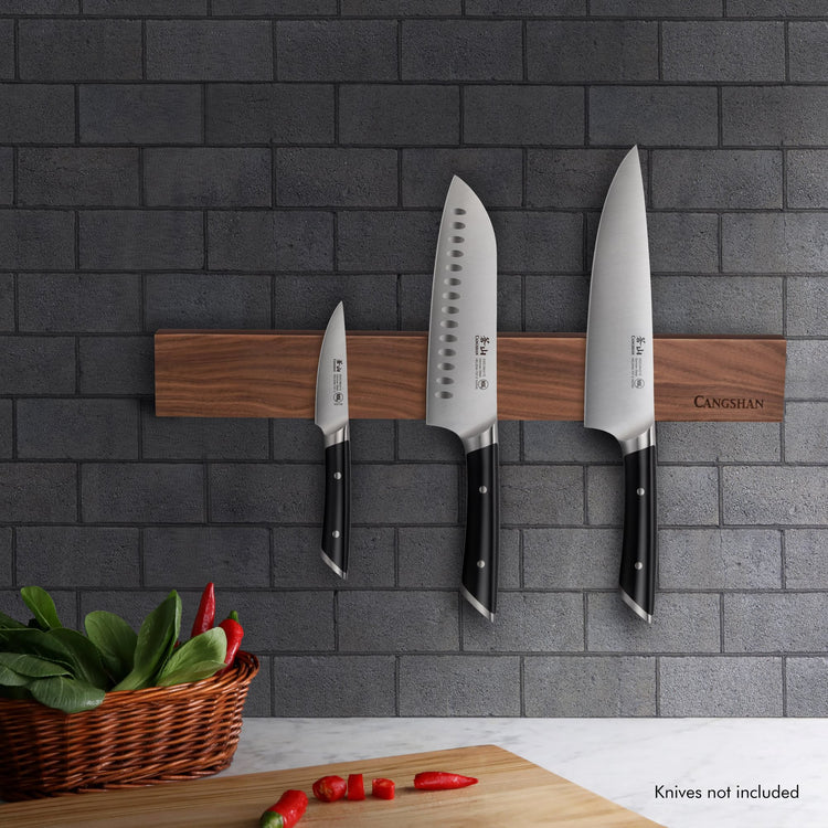 Walnut magnetic knife bar mounted on gray brick wall holding three knives with black handles above a cutting board and basket