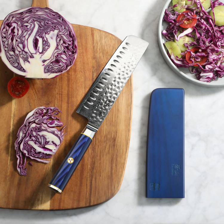 Nakiri knife with hammered blade and blue handle on wooden board, sliced purple cabbage, and blue knife sheath nearby.