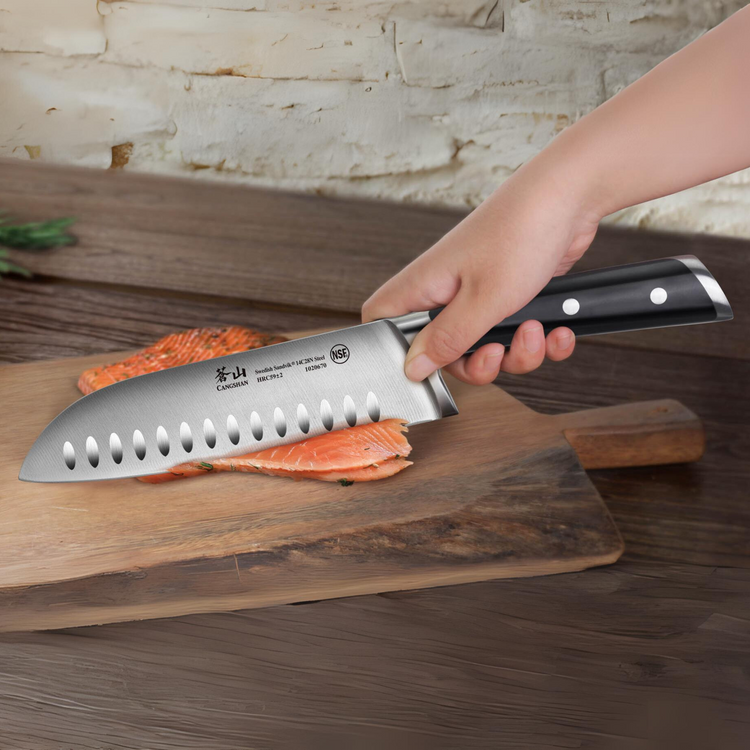 Hand holding a black-handled chef's knife slicing a piece of salmon on a wooden cutting board.