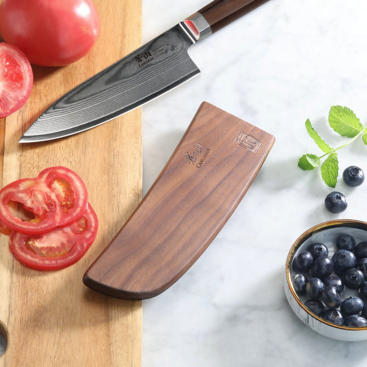 Walnut magnetic knife sheath next to a chef knife, sliced tomatoes on a wooden board, and a bowl of blueberries on a marble s