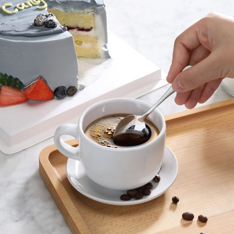 Hand holding a stainless steel espresso spoon stirring black coffee in a white cup on a wooden tray, with a sliced cake and b