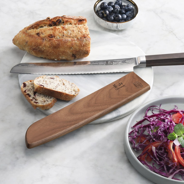 Walnut magnetic knife sheath covering a serrated knife on a marble surface next to bread, blueberries, and a salad bowl.