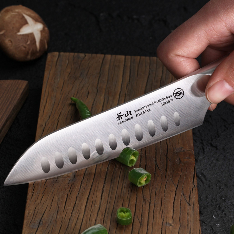 Hand holding a 5-inch Santoku knife with hollow edge, slicing green chili on a wooden cutting board.