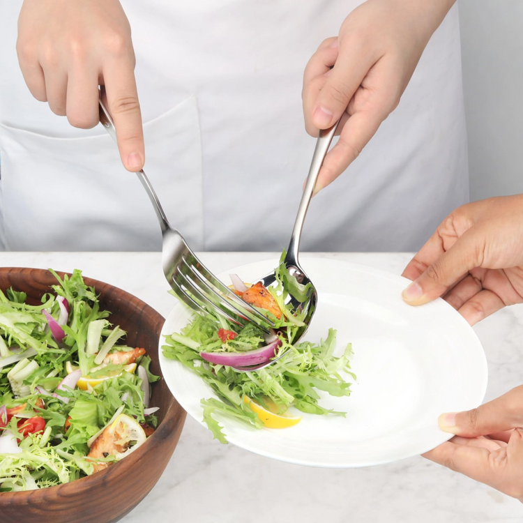 Hands using stainless steel serving fork and spoon to transfer salad from wooden bowl to white plate.