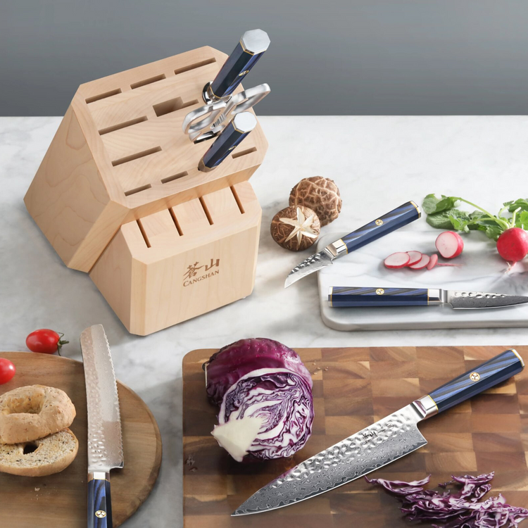 Maple knife block with two knives and kitchen scissors inserted, surrounded by vegetables and additional knives on a countert