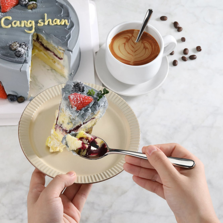 Slice of layered cake with gray frosting and strawberry on a plate, a cup of coffee, and a hand holding a teaspoon.