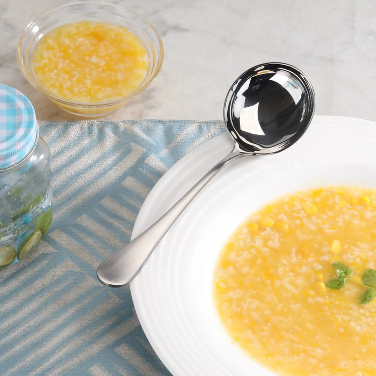Stainless steel soup ladle with matte finish handle resting on a white bowl of yellow soup.