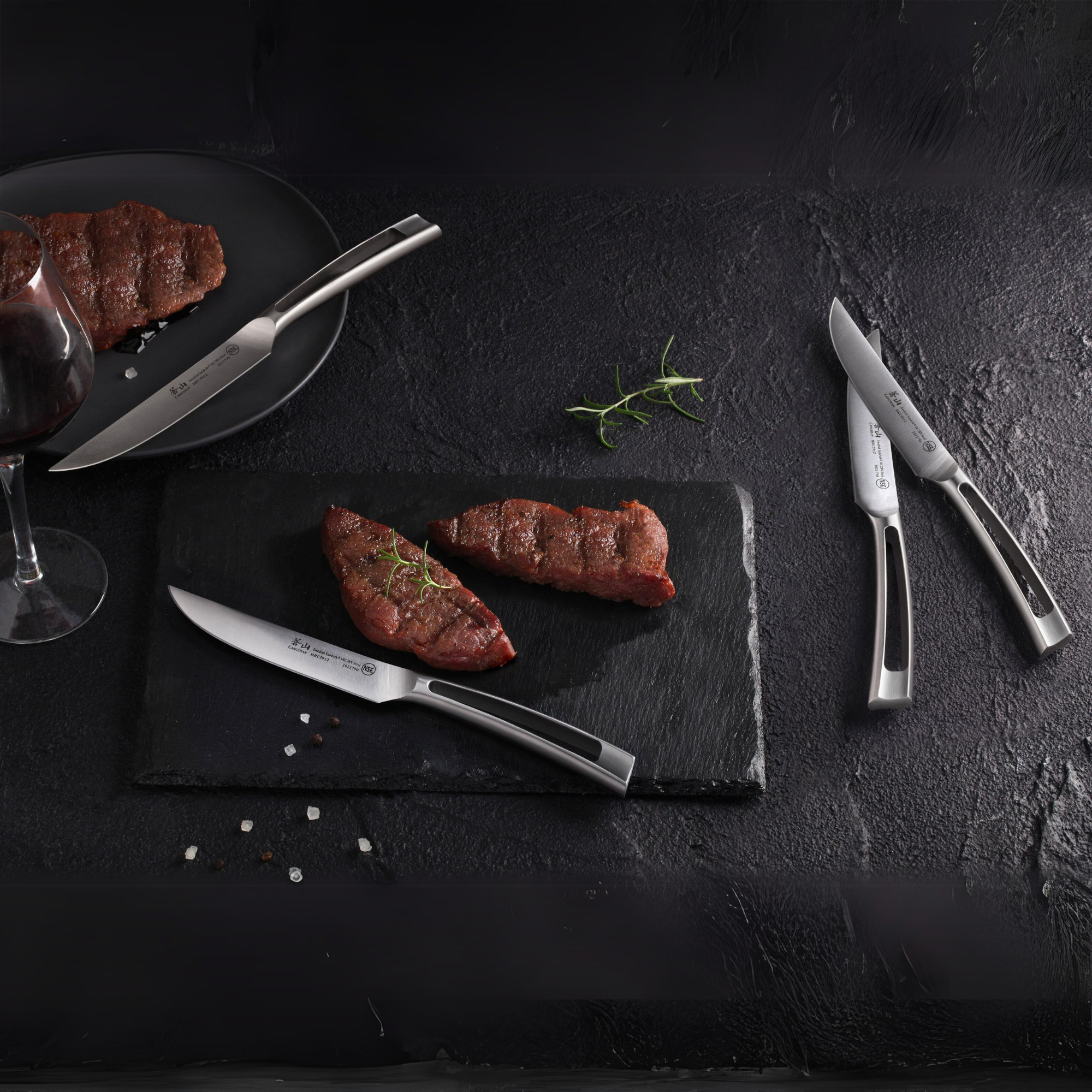 Four stainless steel steak knives with black handles displayed with cooked steak pieces on a black surface.