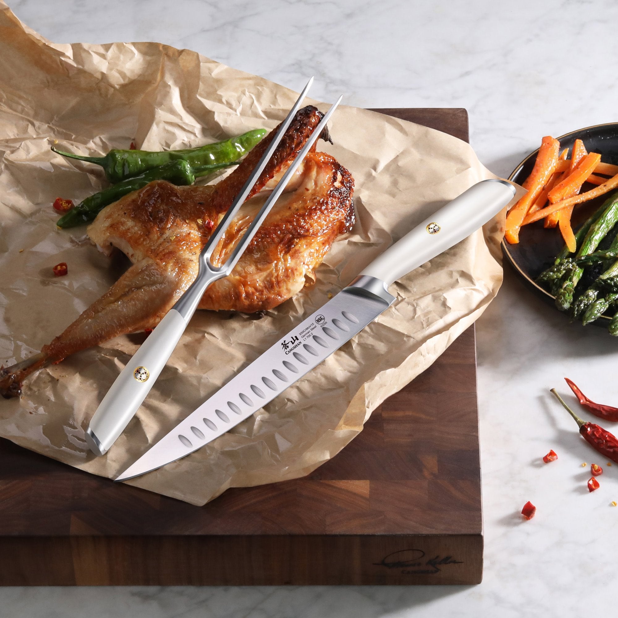 Carving fork and knife with white handles on parchment paper next to roasted chicken and green chili peppers.