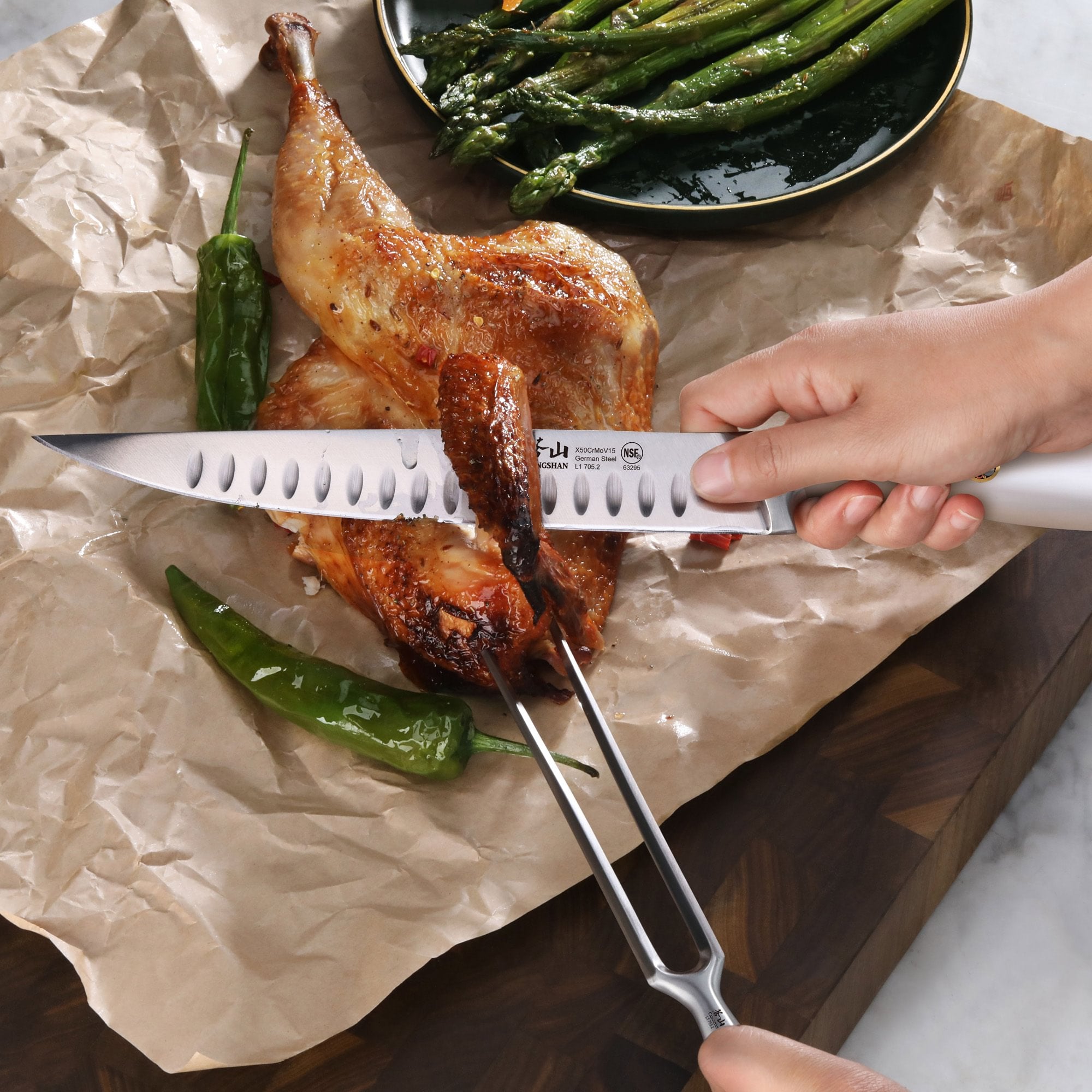 Hand holding a carving knife slicing a roasted chicken leg held by a carving fork on brown parchment paper with green peppers