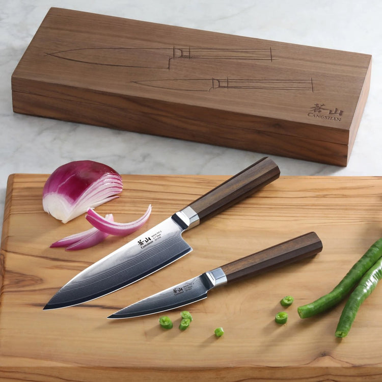 Two knives with dark wooden handles on a wooden cutting board with sliced onion and green chili, next to a wooden box.