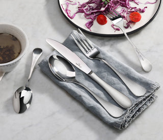 
                    
                      Stainless steel fork, knife, and spoon with matte finish handles on a gray napkin, next to a plate with salad and a cup.
                    
                  