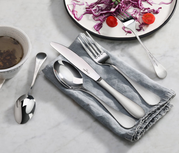 Stainless steel fork, knife, and spoon with matte finish handles on a gray napkin, next to a plate with salad and a cup.