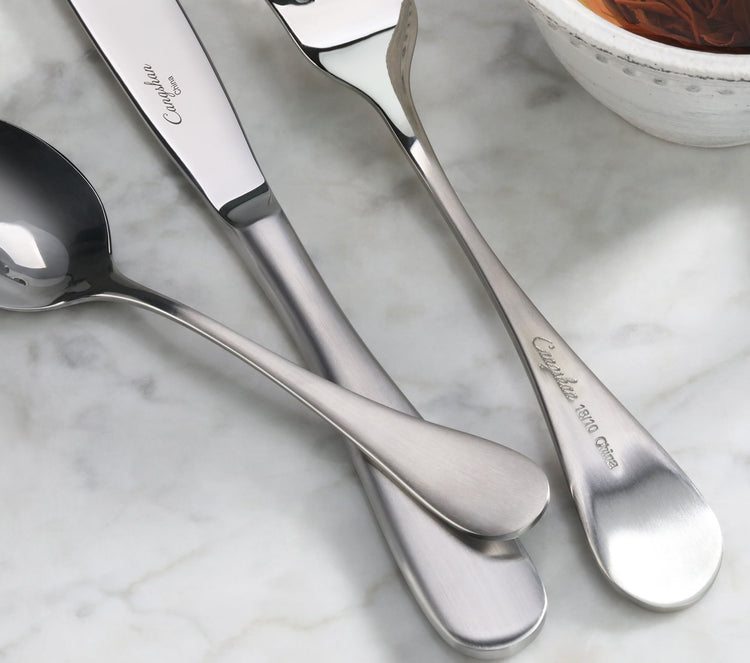 Three stainless steel flatware pieces with matte finish handles on a marble surface near a bowl.