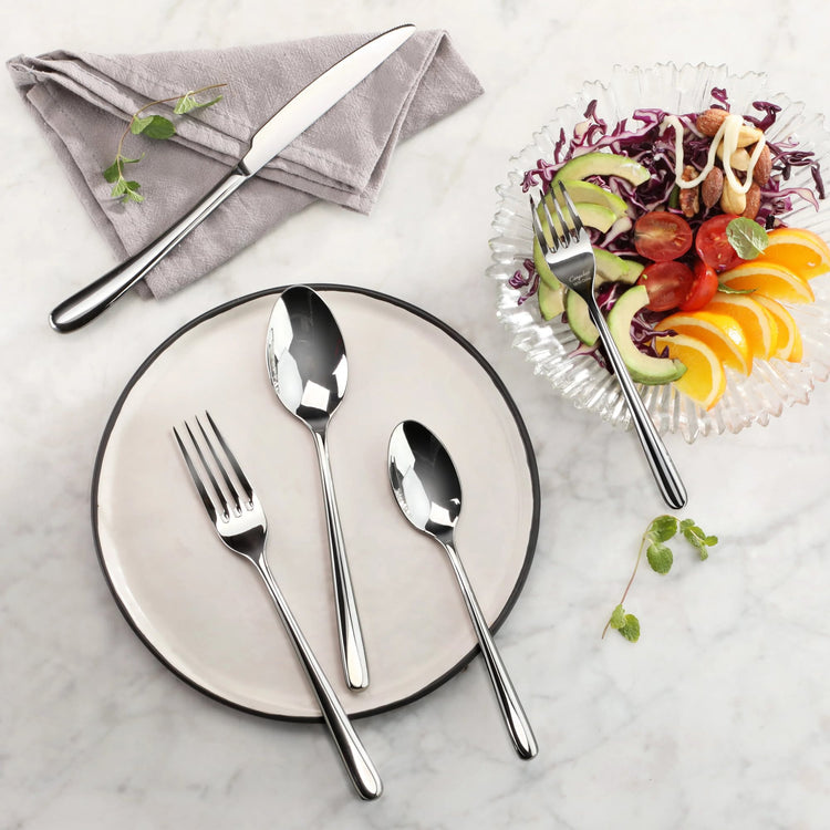 Set of polished stainless steel fork, knife, and spoons arranged on a plate and napkin beside a salad bowl.