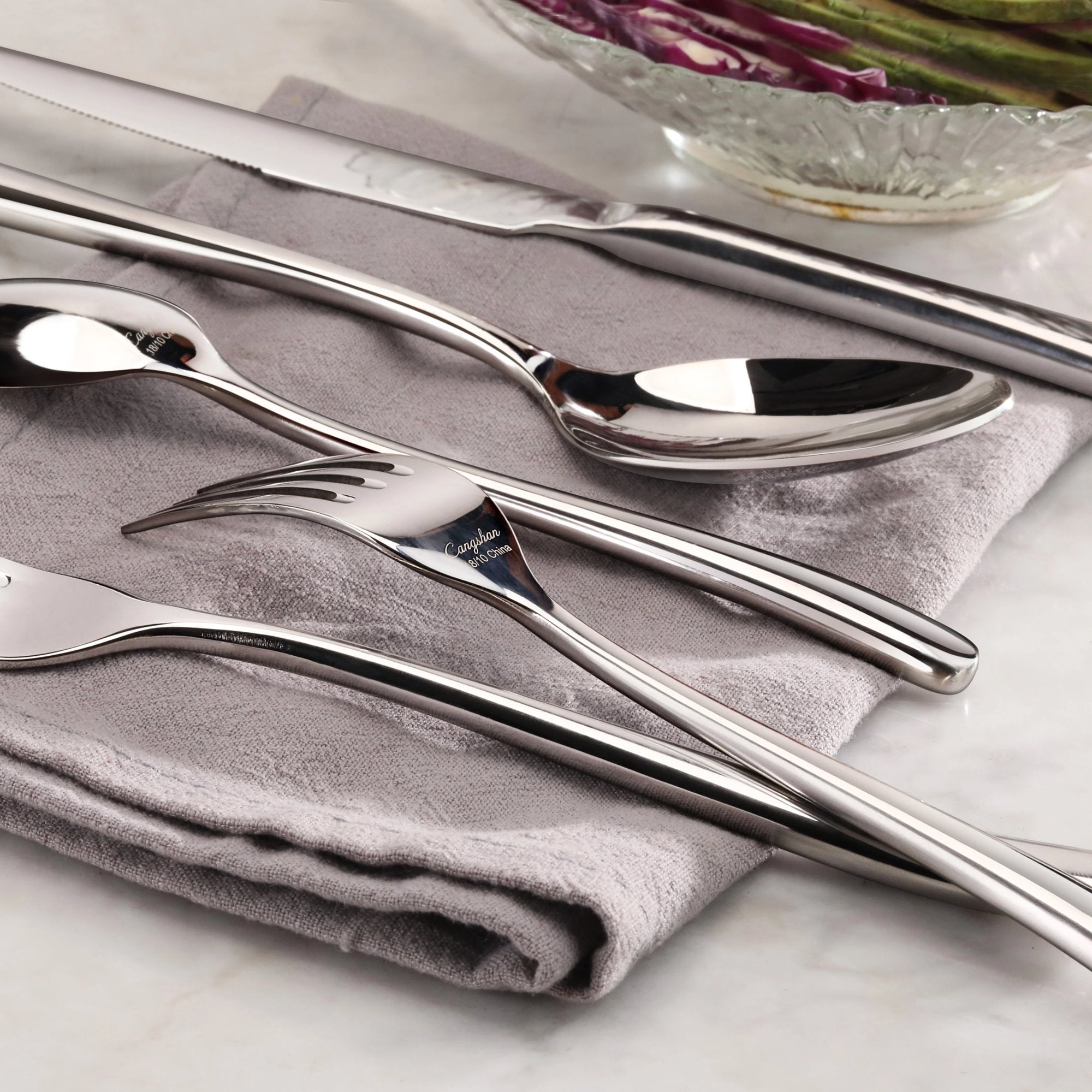 Four pieces of polished stainless steel flatware—a knife, spoon, fork, and teaspoon—rest on a folded gray cloth.
