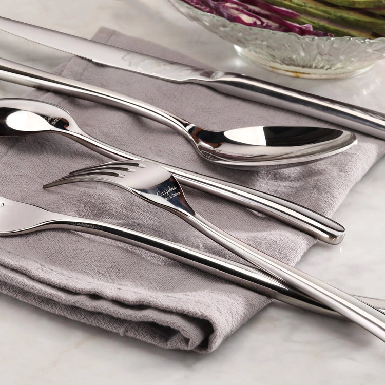 Four pieces of polished stainless steel flatware—a knife, spoon, fork, and teaspoon—rest on a folded gray cloth.