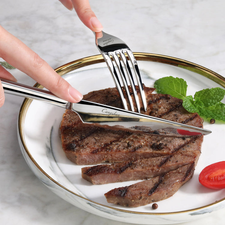 Hand holding a fork and knife cutting a grilled steak on a white plate with gold rim, garnished with mint and cherry tomato.