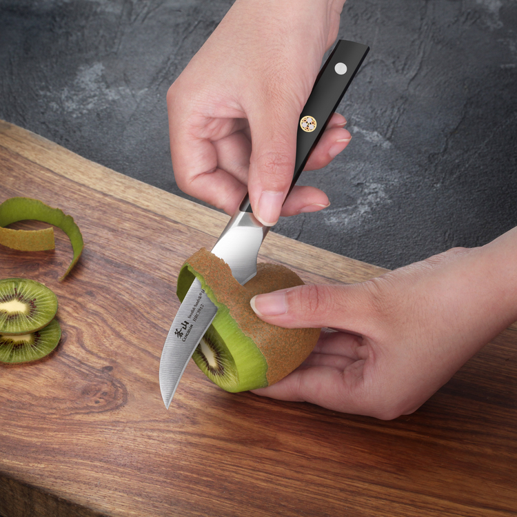 Hands peeling a kiwi with a small curved knife over a wooden cutting board.