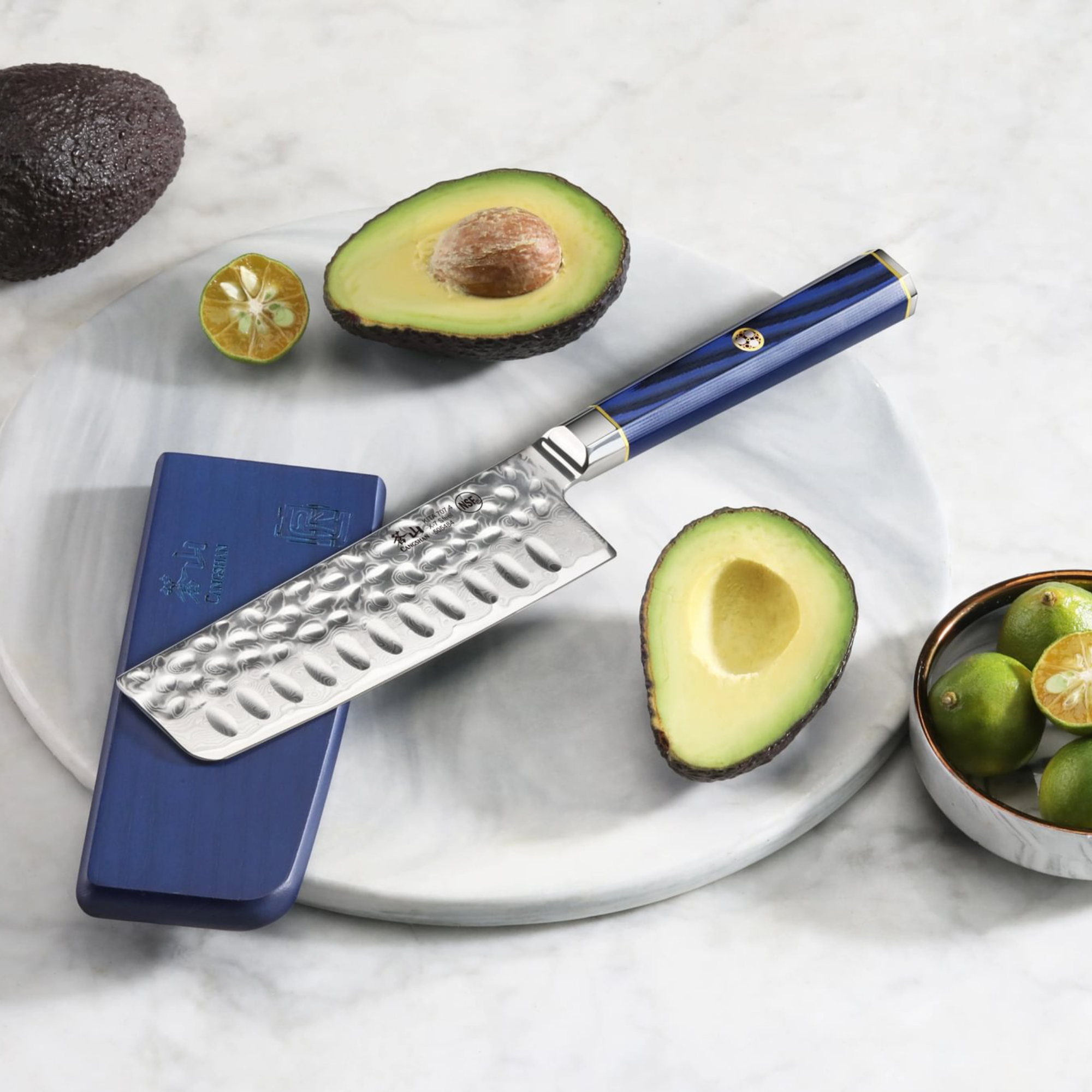 Nakiri knife with hammered blade and blue handle on marble board with halved avocado and calamansi fruit.