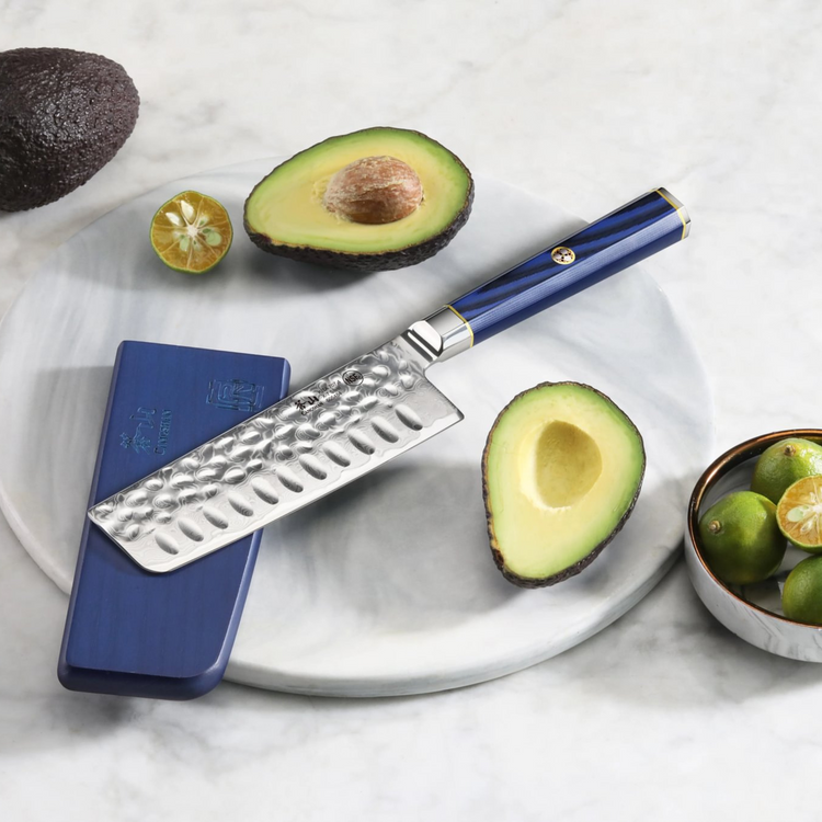 Nakiri knife with hammered blade and blue handle on marble board with halved avocado and calamansi fruit.