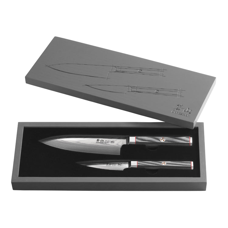 Two Damascus steel knives with black handles displayed in an open gray box with knife illustrations on the lid.