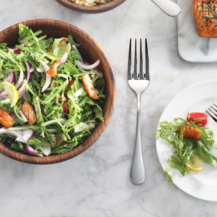 Serving fork with matte finish handles placed on a marble surface next to a wooden bowl of salad and a white plate with salad