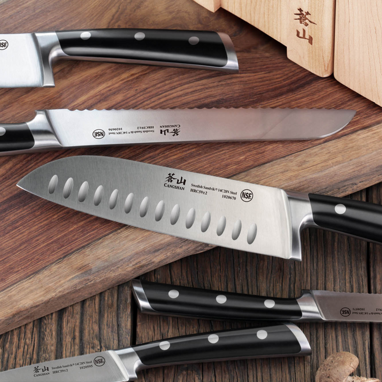 Set of forged stainless steel kitchen knives with black handles displayed on wooden surface near a wooden block.