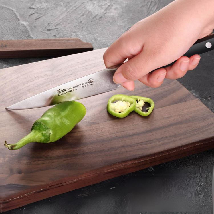 Hand holding a knife slicing a green chili pepper on a wooden cutting board.