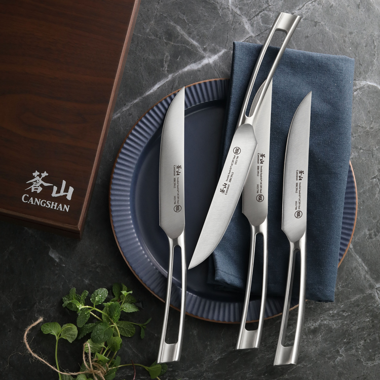 Four stainless steel steak knives with hollow handles arranged on a blue plate and napkin, next to a wooden knife box.