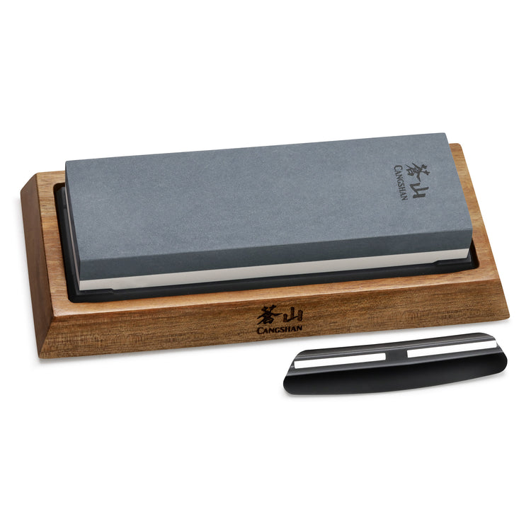 Rectangular dual-sided whetstone with gray and white sharpening surfaces on a wooden base, next to a black and silver knife h