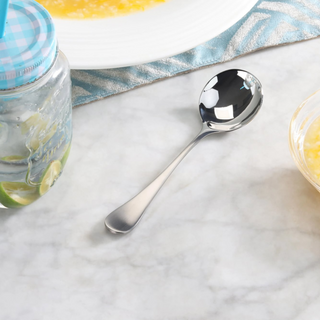 
                    
                      Stainless steel bouillon spoon with matte finish handle resting on a marble surface near a jar and a bowl.
                    
                  
